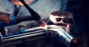 A welder in a mask with welding tools using the TIG welding method to conjoin two stainless steel pipe parts.