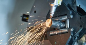 A laser tube cutting machine cutting into a metal pipe with orange sparks flying off the instrument and pipe.