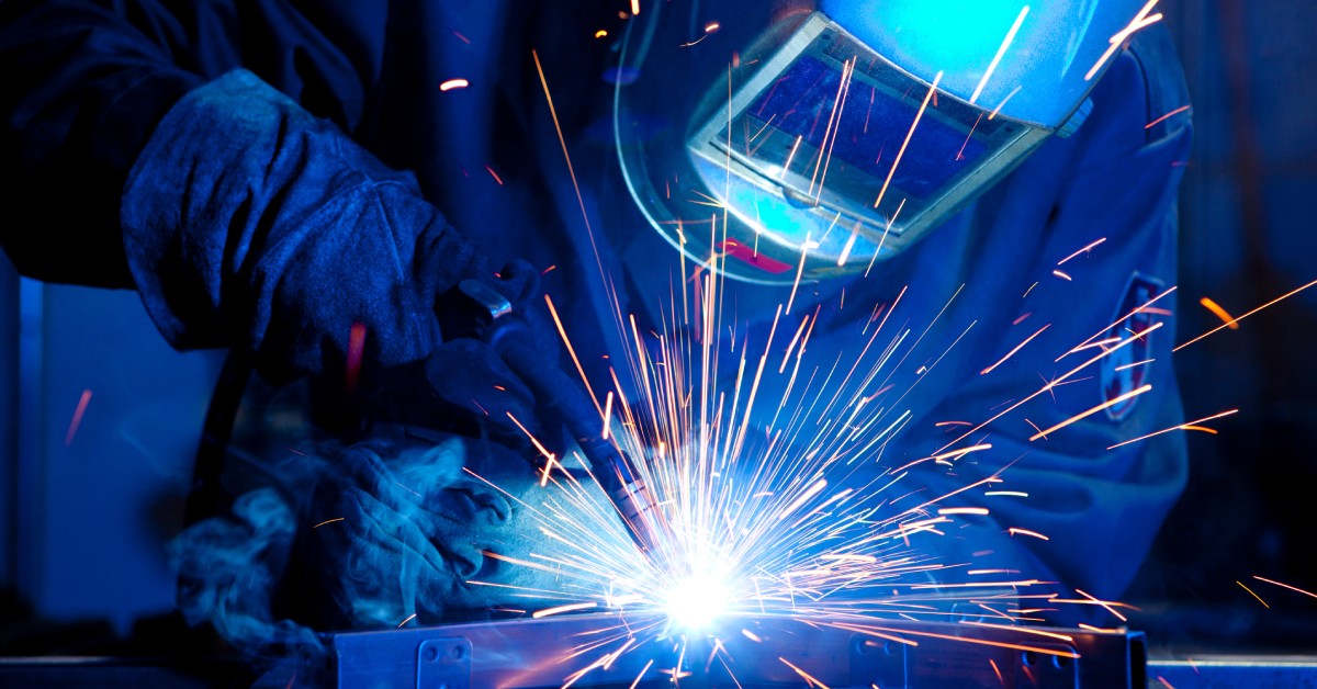 A welder in a dark, blue lit room, wearing a welder's mask. They weld a rectangular piece of metal onto a metal disc.