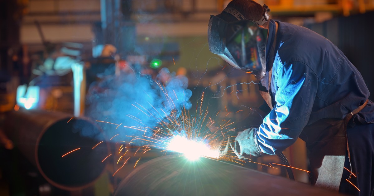 A welder wearing a mask and apron in an industrial setting. They lean over a large metal pipe and weld it with their torch.