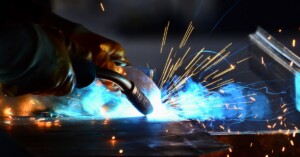 A close up of a welder's hands, torch welding two pieces of metal together, causing sparks and blue flames.