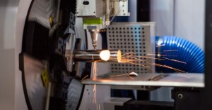 A tube laser cutting machine cutting segments out of a metal pipe. There are orange sparks flying from the cutting site.