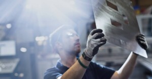 An industrial worker wearing gloves holds up a thin sheet of metal with squares cut into it for inspection.