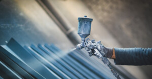 An industrial worker wearing grey gloves and a shirt holding a grey paint gun spraying grey paint mist over metal plates.