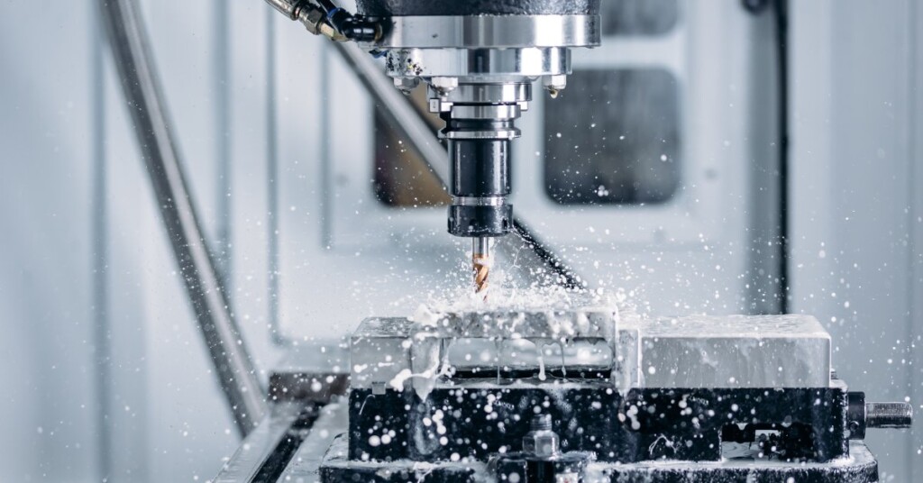 A side profile of a CNC machine drilling a bronze drill bit into a stationary block of metal with splashing water.