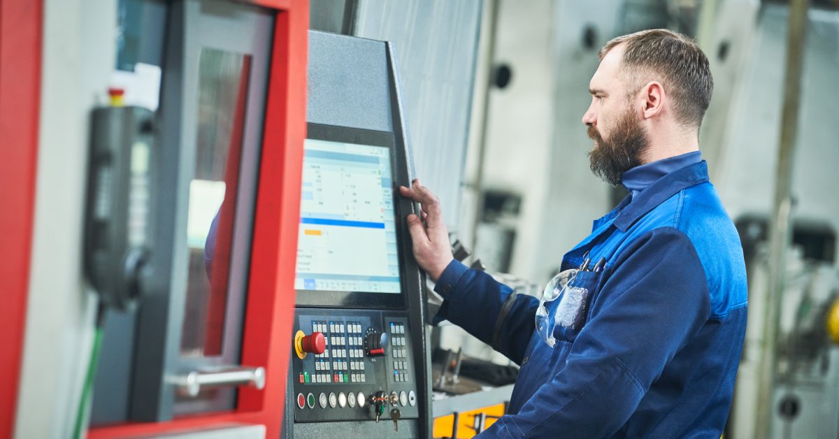 A male industrial worker in a blue jumpsuit observes and operates the main functioning terminal of a machine.