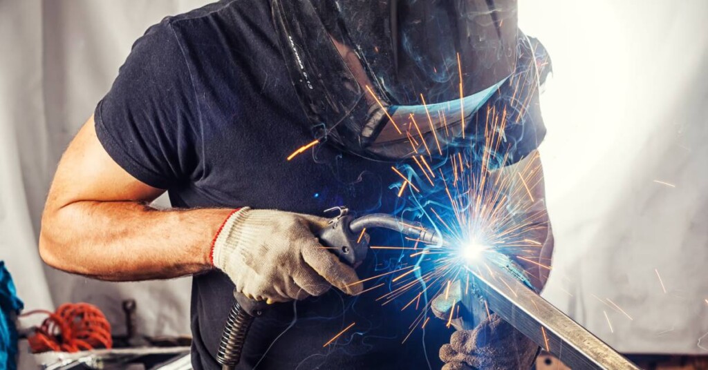 A man wearing a welder's mask, a welder's gloves, and a T-shirt uses a welding torch on a rectangular metal beam.