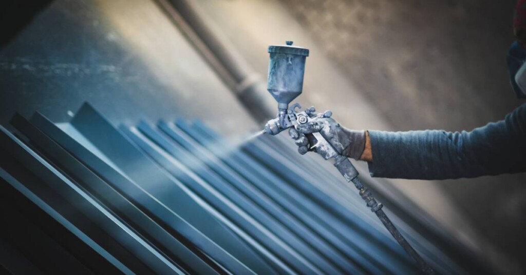 A person wearing a brown glove uses a spray gun to coat the surface of metal sheets placed against a wall in a row.