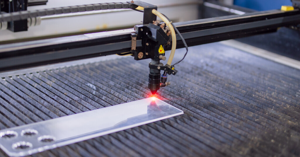 A laser-cutting device is firing a red beam at the surface of a glass sheet with four precise, circular holes on one side.