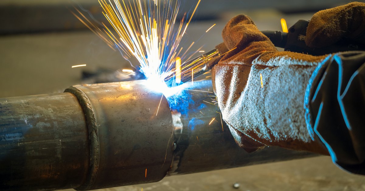 A person wearing thick brown gloves uses an arc welder to close the gap between two section of a large pipe.