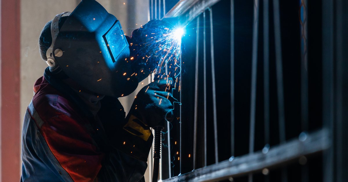 A person wearing a welding mask, gloves, and thick clothing using an arc welder on a surface with metal bars and beams.